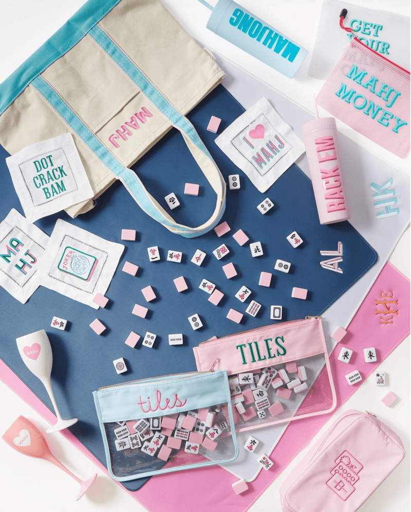 Flat lay featuring a cream and teal embroidered Mahjong canvas tote with “MAHJ” stitched in pink, surrounded by Mahjong-themed accessories including embroidered cocktail napkins, pouches labeled “TILES,” Mahjong tiles, tumblers, and personalized drinkware for a playful and stylish game night setup.