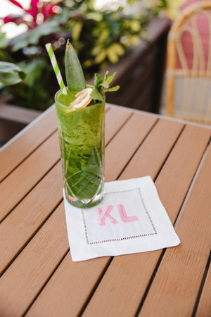 A cocktail napkin is an embroidered with a pink monogram and placed under a green drink.