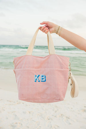A person at the beach holds up a pink striped cooler tote with a blue monogram.