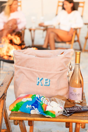 A pink monogrammed cooler tote is customized with a blue monogram and placed by a campfire with supplies for smores