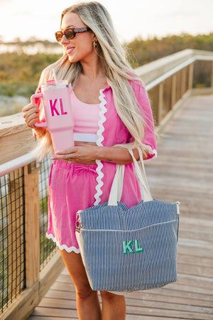 A girl on a board walk holds a blue cooler tote bag with a green monogram and a pink 40 oz tumbler with a pink monogram.