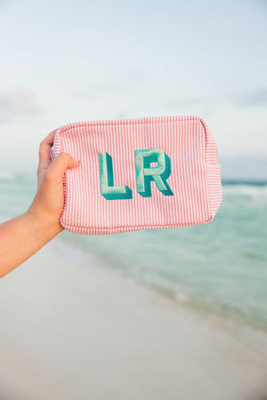 A person holds up a pink seersucker pouch with a mint and turquoise monogram at the beach.