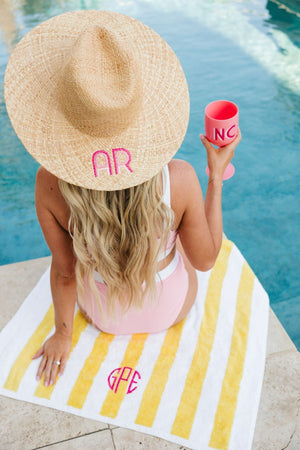 A girl sits on the edge of a pool wearing an embroidered straw hat with a pink monogram and holds a pink wine glass with a pink monogram.