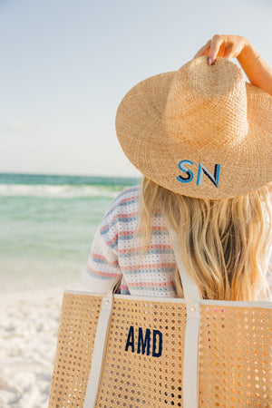 A woman at the beach holds onto her straw hat which has been embroidered with a blue monogram and holds a cane tote with a navy monogram.