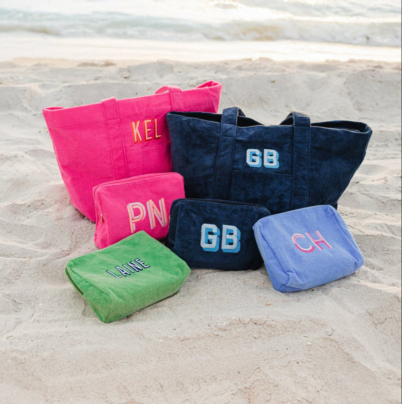 A stylish set of personalized terry cloth tote bags and matching cosmetic pouches displayed on a sandy beach with ocean waves in the background. The set includes a hot pink tote embroidered with "KEL", a navy tote with "GB", and multiple coordinating cosmetic pouches in pink, green, navy, and blue with custom monograms like "PN," "GB," "LANE," and "CH." These soft, plush bags from Sprinkled with Pink are perfect for beach days, vacations, and personalized gifting, offering a chic and functional way to carry