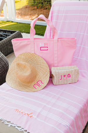 A pink coated canvas tote, straw pouch and a straw hat are placed on a beach chair covered by a pink Turkish towel with an orange script name embroidered on it.