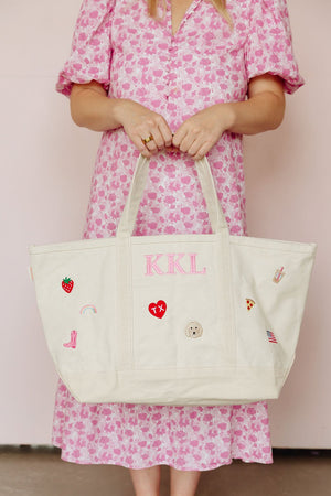Woman in a pink floral dress holding a natural canvas tote monogrammed 'KKL' in pink, decorated with strawberry, rainbow, cowboy boot, heart, and dog icon patches