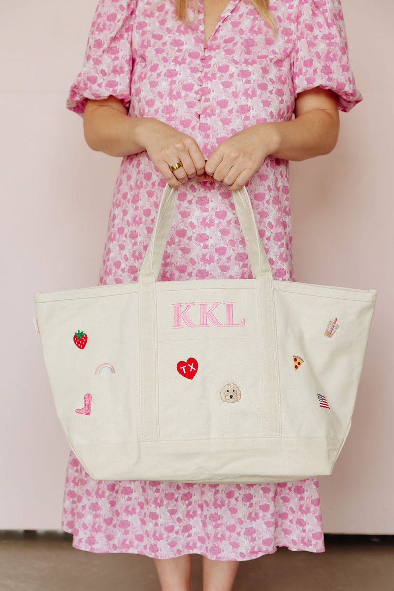 Woman in a pink floral dress holding a natural canvas tote monogrammed 'KKL' in pink, decorated with strawberry, rainbow, cowboy boot, heart, and dog icon patches