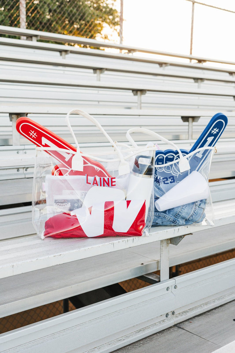 Gameday Clear Tote Bags rest on bleachers, red and blue kits with matching pompoms and bold name decals.