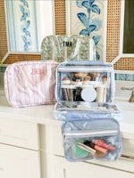 Open blue hanging toiletry bag filled with makeup brushes and compact, beside a pink striped pouch monogrammed JMH and a green floral stripe bag monogrammed TMA on a bathroom counter