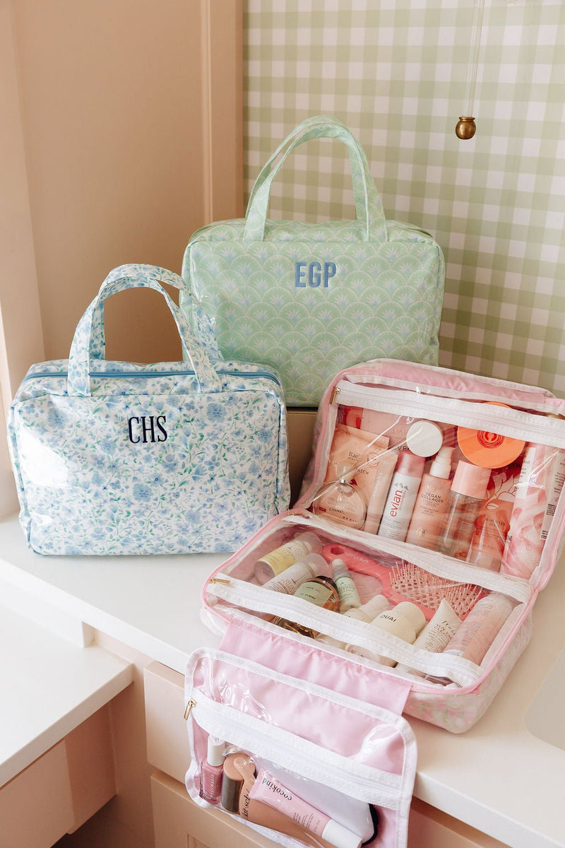 Sage green scalloped hanging toiletry bag monogrammed EGP and a blue floral hanging toiletry bag monogrammed CHS, beside an open pink striped hanging organizer filled with toiletries on a bathroom counter