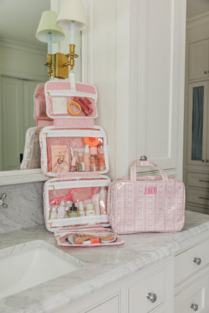 Pink monogrammed hanging toiletry bag unfolded on bathroom counter, showcasing multiple compartments and travel essentials.