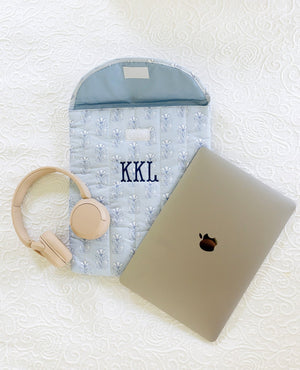 A personalized light blue quilted laptop sleeve with 'KKL' embroidered in navy, styled on a white textured blanket alongside a closed silver laptop, beige over-ear headphones, and a matching beige pouch.