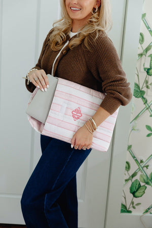 A woman wearing a brown cardigan and dark blue jeans holds a pink and white striped tote bag with a pink heart design and a gray shoulder strap. The background shows a white door and a green botanical patterned panel.