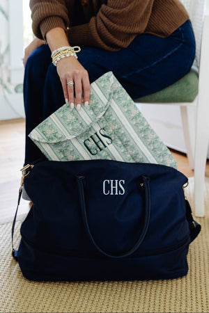 A navy blue personalized duffel bag with white monogrammed initials 'CHS' sits on a woven rug in a home setting, with a person sitting on a chair partially visible behind it placing a green patterned envelope or mailer into the bag.