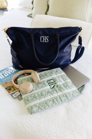 A navy blue monogram duffel bag with gold hardware sits on a white textured bed, styled with a green floral-patterned laptop sleeve, beige headphones, and a closed silver laptop.