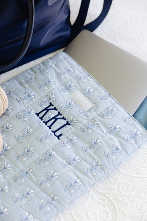 A light blue quilted monogrammed laptop sleeve with a blue floral pattern and the monogram 'KCL' embroidered in navy, styled on a white textured blanket next to a navy tote bag and a silver laptop.