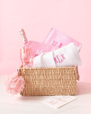 A basket is filled with a white bath robe, pink wine glasses, and pink roadies.