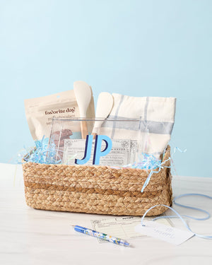 A basket is filled with an acrylic box used as a recipe box, a kitchen towel, a spatula, and some sweet treats.