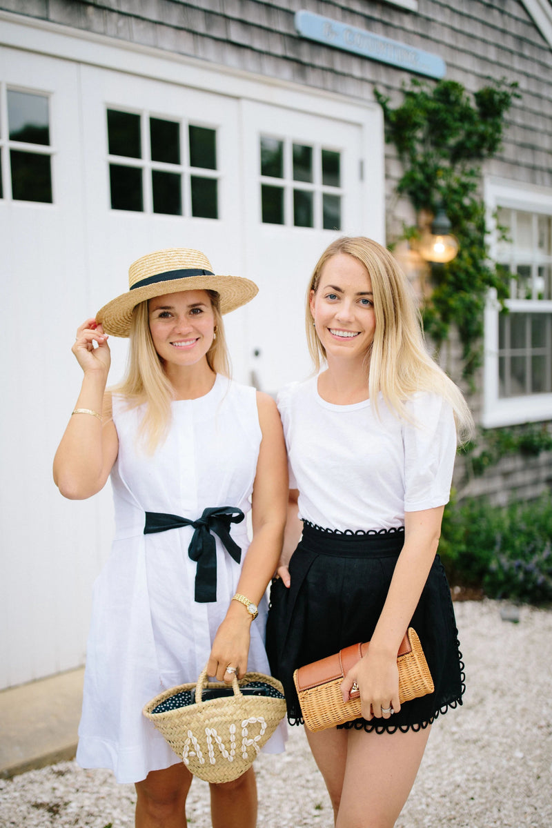 Two women dressed in classic black-and-white summer outfits pose stylishly outside a coastal cottage. The woman on the left accessorizes with a Monogrammed Boater Hat featuring a black ribbon and initials, adding a timeless, personalized touch to her breezy, preppy look.
