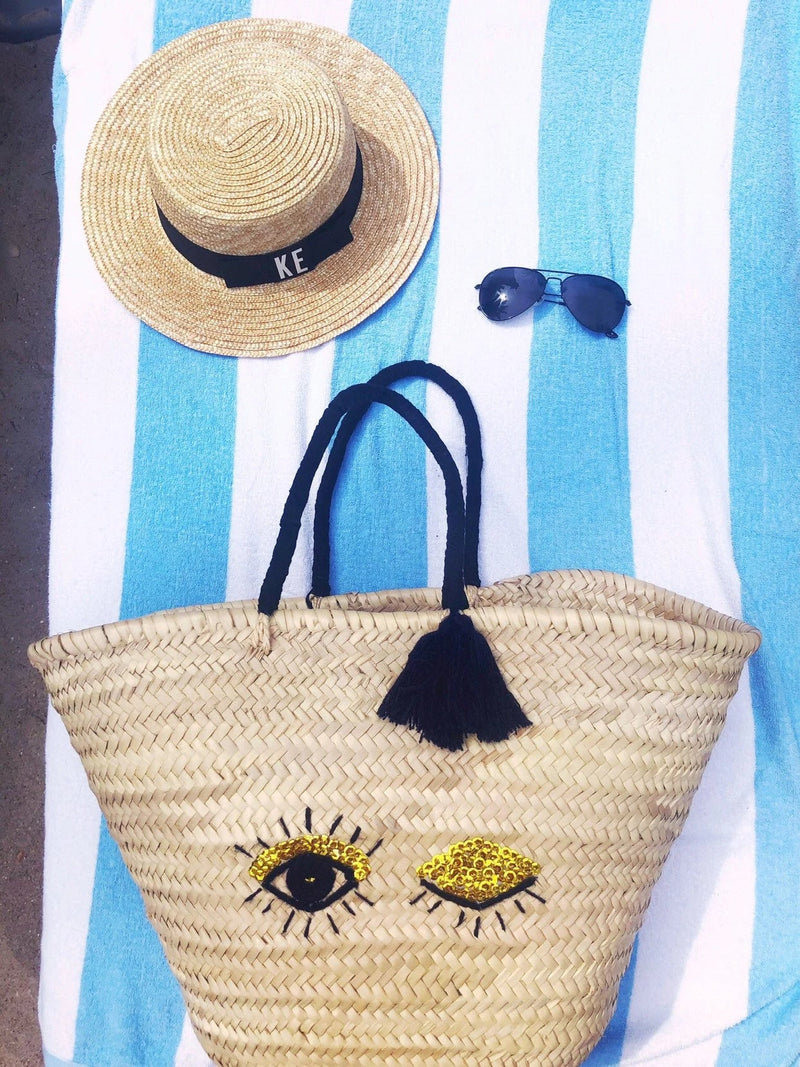 Flat lay of a beach setup featuring a Monogrammed Boater Hat with black ribbon and white "KE" initials, paired with black aviator sunglasses and a woven straw beach tote adorned with playful sequin eye and lemon designs—all arranged on a blue and white striped towel for a chic coastal vibe.
