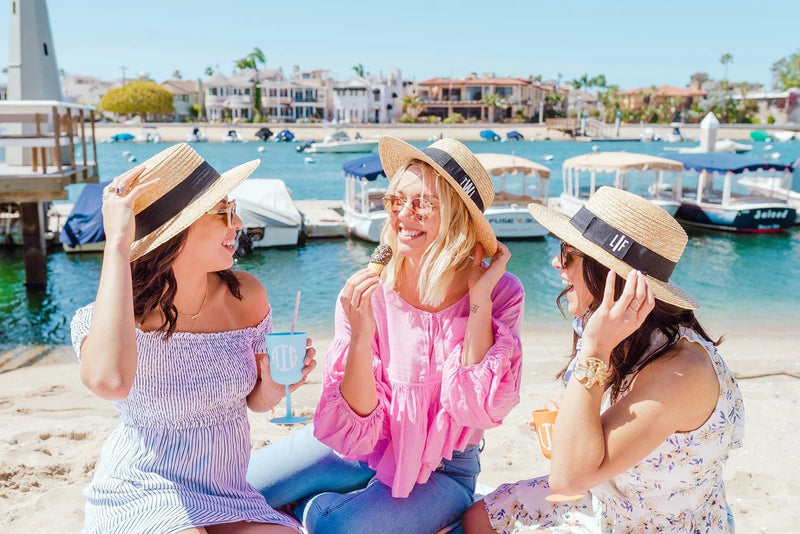 Three women enjoying a sunny beach day wearing Monogrammed Boater Hats with personalized initials on black hatbands, sipping from monogrammed tumblers and laughing by the water with docked boats and waterfront homes in the background.