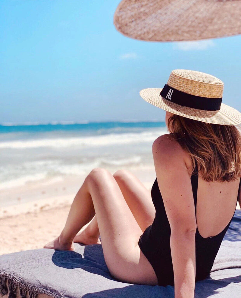 Woman lounging on a beach chair in a black swimsuit, wearing a personalized Monogrammed Boater Hat with “AL” embroidered on a black band, capturing effortless seaside elegance under the sun.