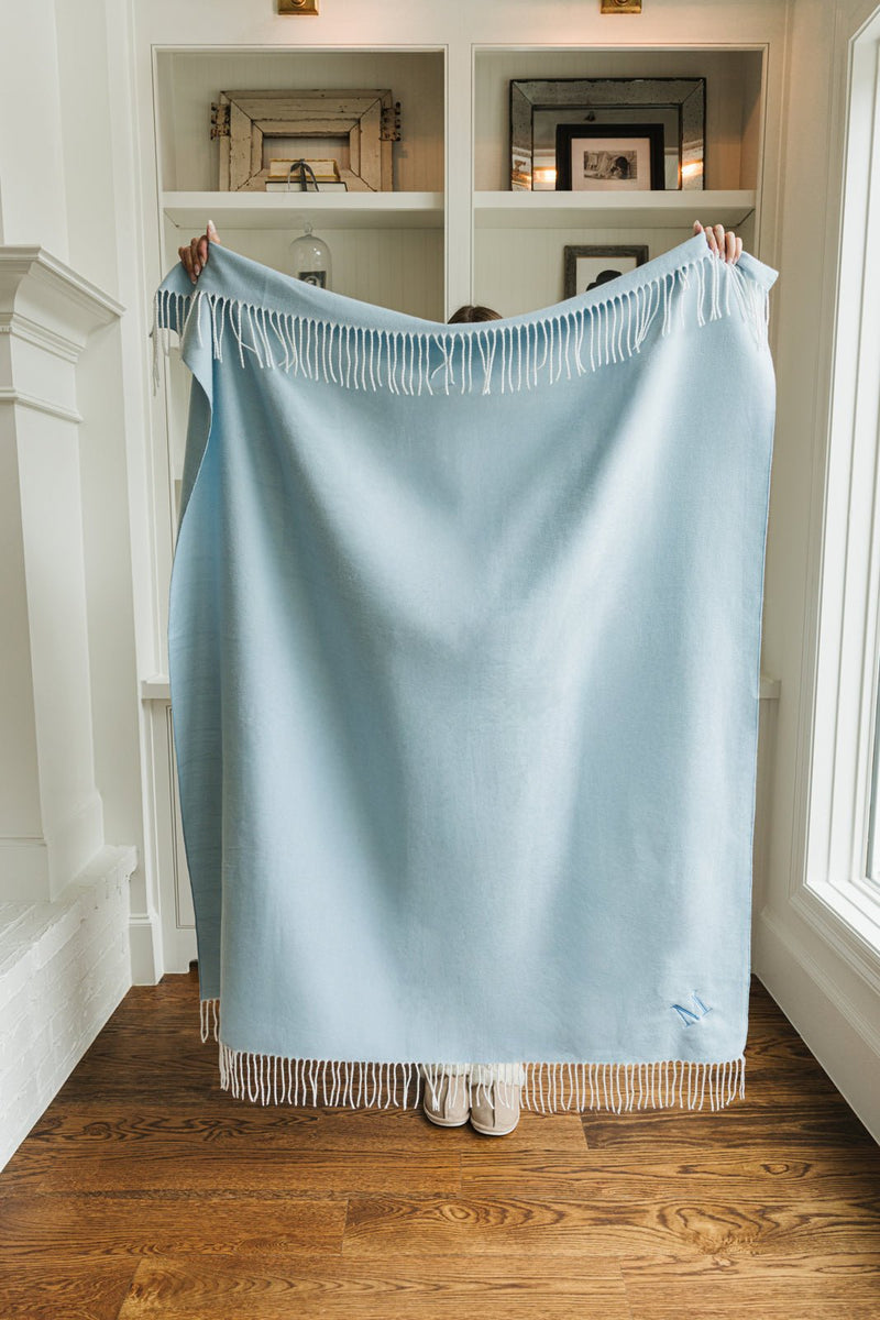 Full view of light blue Monogrammed Woven Blanket with fringe detailing held up to show size.