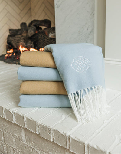 Stack of blue and camel Monogrammed Woven Blankets with embroidered initials by the fireplace.