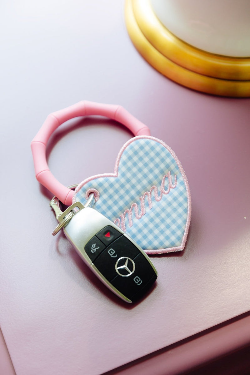 A pink silicone keychain with a heart-shaped bag tag featuring a light blue and white gingham pattern and the embroidered name 'Emma', holding a Mercedes-Benz key fob, displayed on a pink night stand.