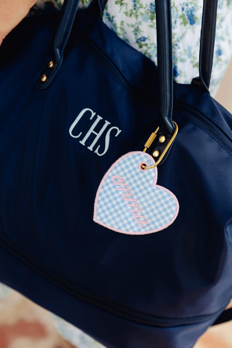A navy blue tote bag with gold-tone hardware, featuring the monogram 'CHS' in white on the front and a heart-shaped light blue gingham key tag with pink script embroidery.
