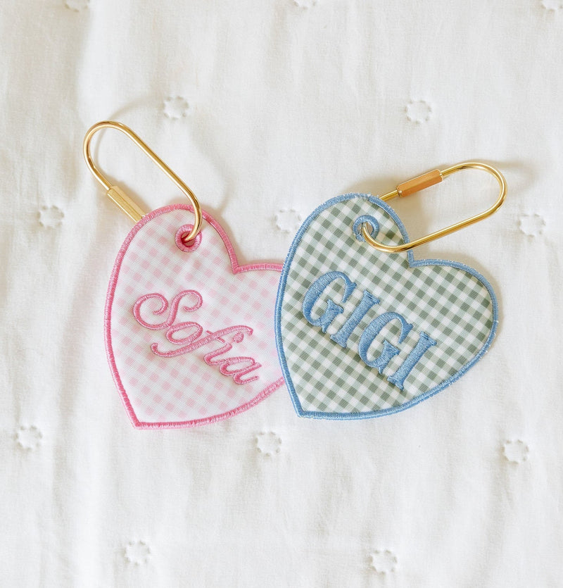 One pink and white gingham heart shaped bag tag next to a green gingham heart shaped bag tag in french blue embroidery in the shape of a heart, y laying on the bed with gold carabeeners.