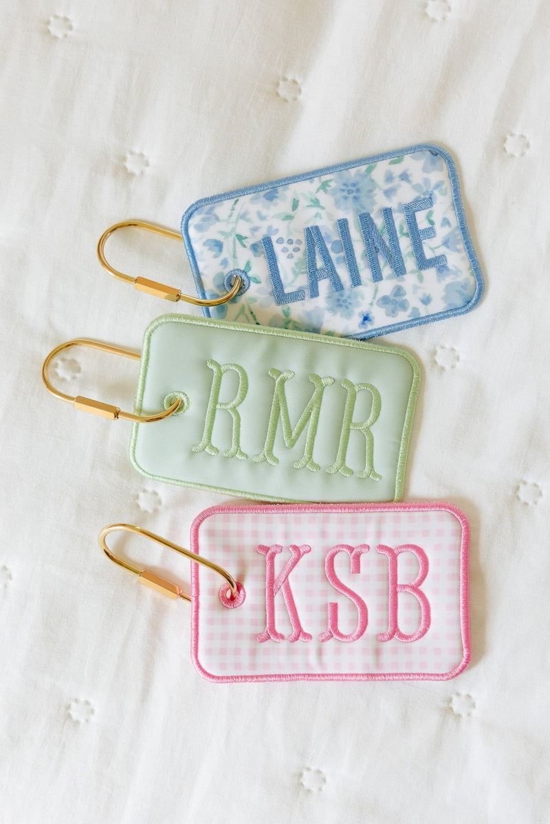 Three rectangular bag tags on gold carabiners: blue floral embroidered 'Laine' in blue, sage green embroidered 'RMR' in green, and pink gingham embroidered 'KSB' in pink.