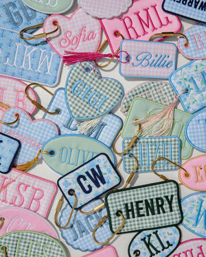A dense flat-lay collection of personalized embroidered name tags in pink, light blue, mint green, and white gingham patterns, each with a gold metal clip and a small tassel.