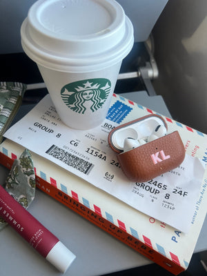 Shadow Monogram AirPods Pro Case in brown leather with pink initials, styled with travel essentials on an airplane tray.