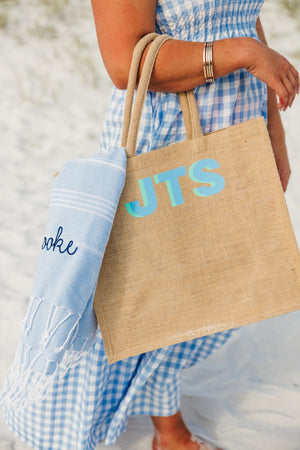 A girl wears a jute bag with a blue monogram on her arm with a blue embroidered towel hanging out of it.