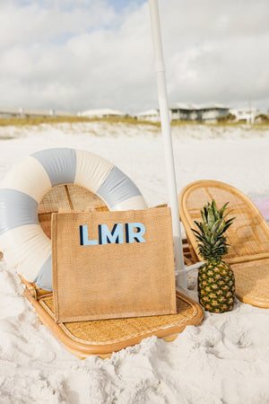 A jute carryall with a blue monogram is placed on a chair at the beach.