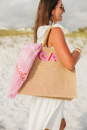 A girl wears a jute bag with a pink monogram on her shoulder with a pink embroidered towel hanging out of it.