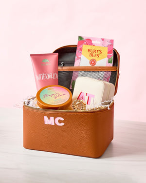 A tan leather train case is filled with with self care products and a pink monogrammed jewelry case