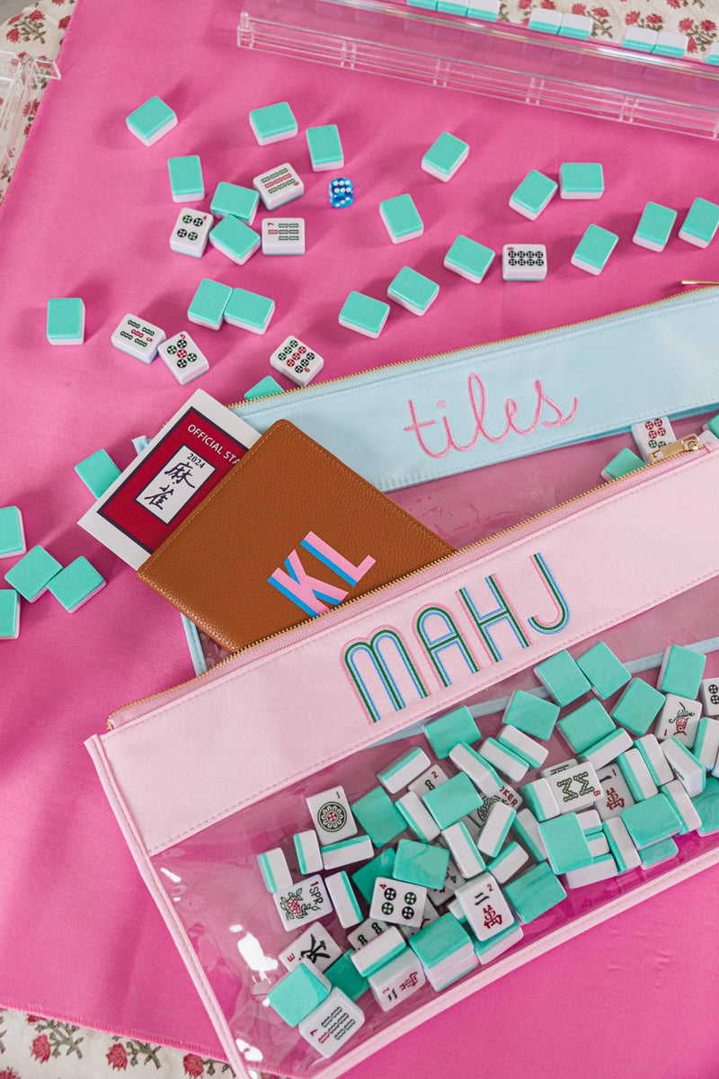 A beautifully styled Mah Jongg set featuring a pink game mat, scattered turquoise and white tiles, and two personalized storage pouches labeled 'tiles' and 'MAHJ.' A tan leather monogrammed cardholder with 'KL' rests on top, alongside an official Mah Jongg rule card, adding a personalized and coordinated touch to the gaming experience.