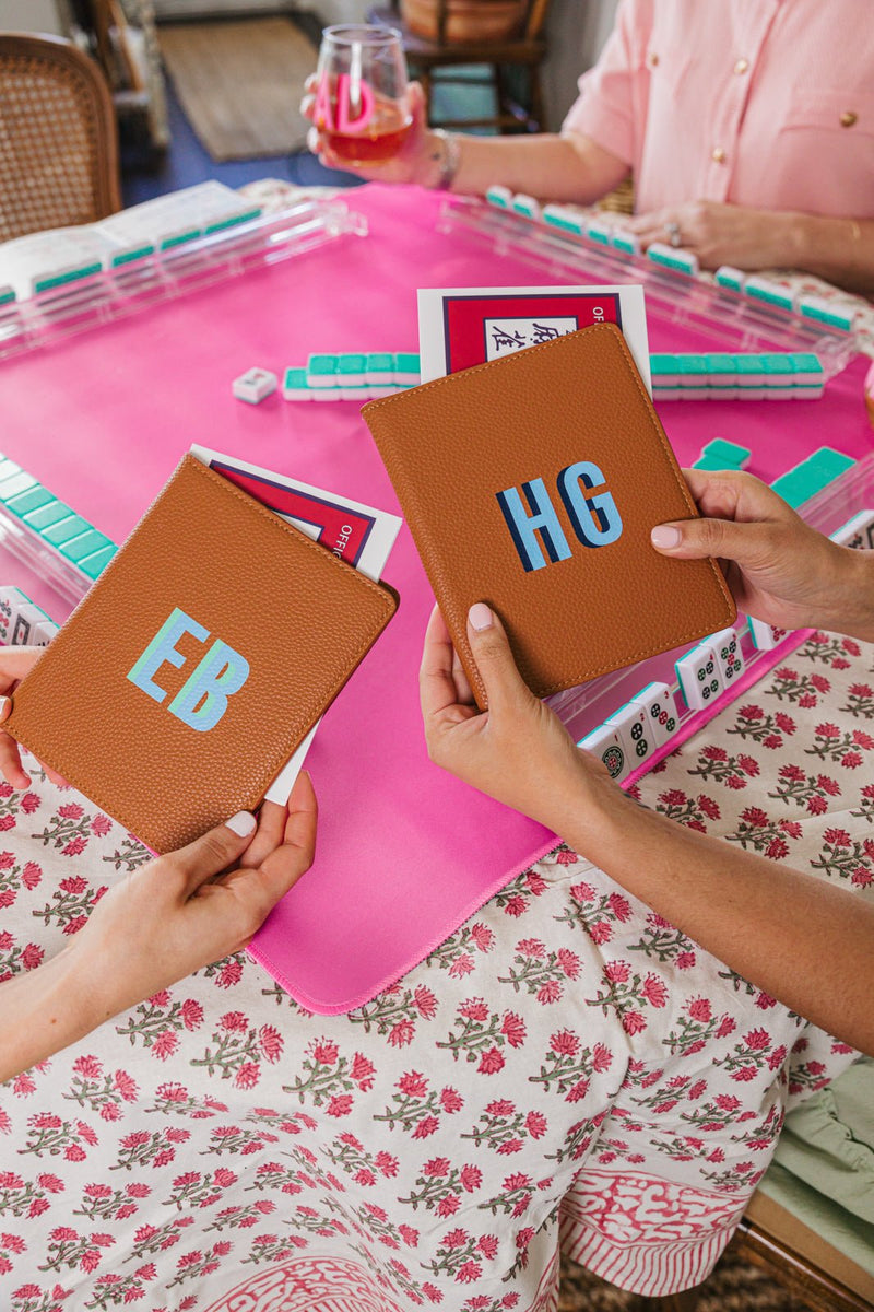 Two hands hold personalized tan leather cardholders, monogrammed with 'EB' and 'HG,' perfectly complementing a chic Mah Jongg setup. The scene features a pink Mah Jongg mat, custom acrylic racks, and a floral tablecloth, creating a coordinated and stylish game night ambiance. The matching set of leather accessories and monogrammed details elevate the experience, making it both functional and fashionable for a personalized Mah Jongg gathering.