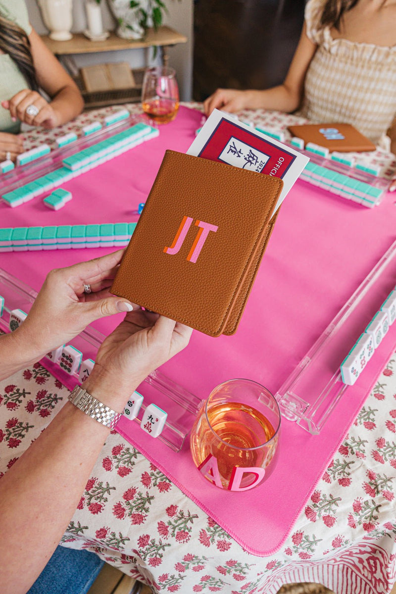 A beautifully curated Mah Jongg setup featuring a personalized tan leather cardholder with the initials 'JT,' holding the 2024 National Mah Jongg League rule card. The game is set on a vibrant pink mat with matching clear tile racks and pastel tiles, creating a stylish and organized gaming experience. A monogrammed wine glass labeled 'AD' adds an extra touch of personalization, while players enjoy the game in the background, showcasing a cohesive and elegant Mah Jongg set.