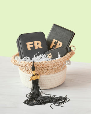 A basket is filled with black leather monogrammed products and a black graduation tassel hangs from the side.