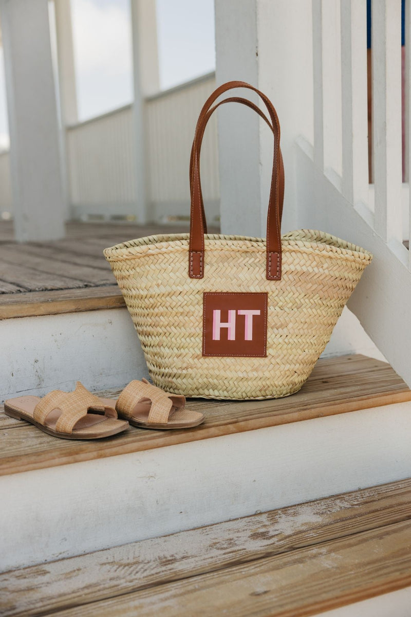 A straw tote with a pink monogram is placed on a dock next to some sandals