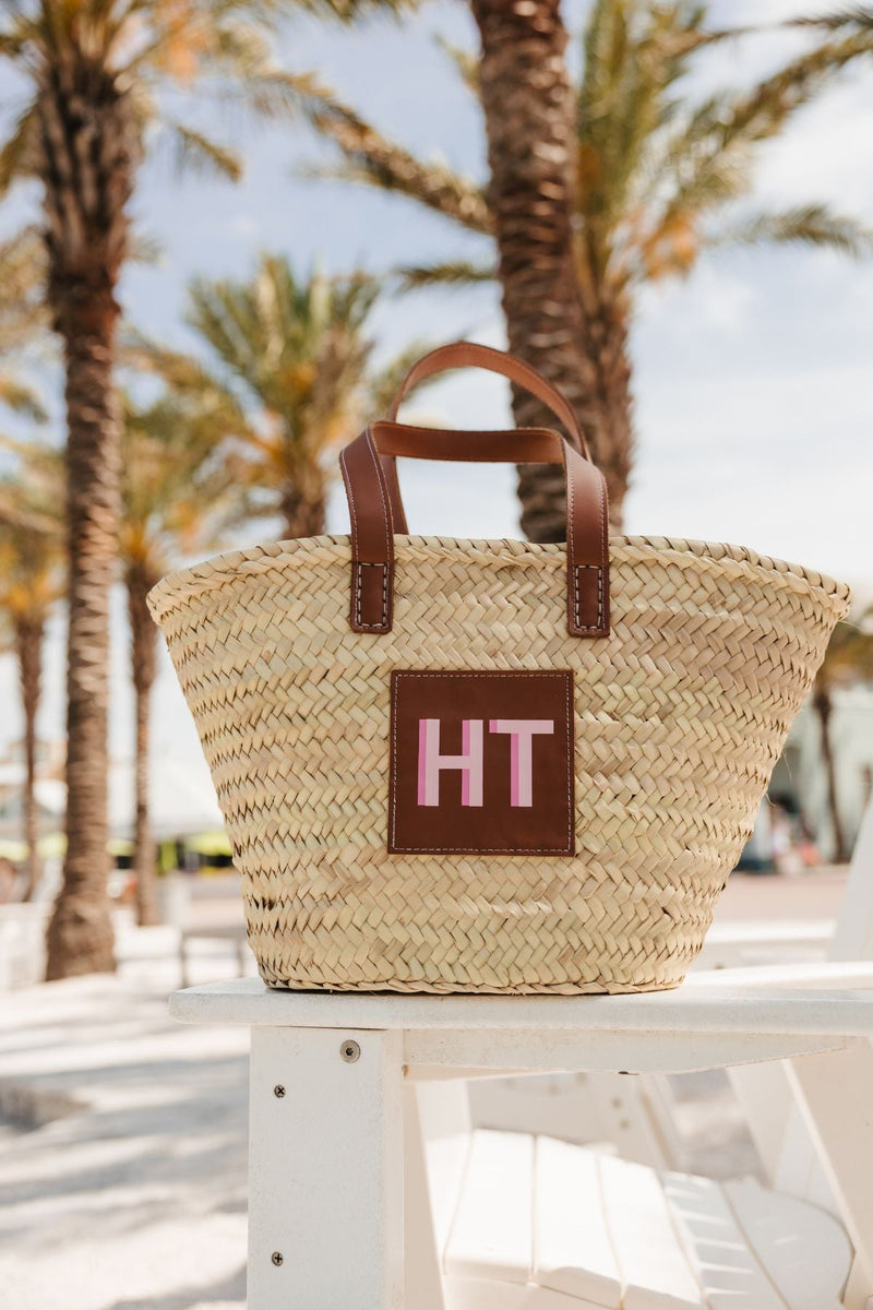 A straw tote sits on a beach chair in front of palm trees with a customized pink monogram