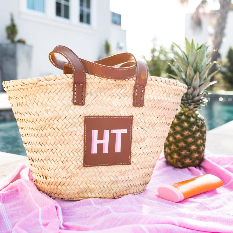 A straw tote bag is customized with a pink monogram at a pool