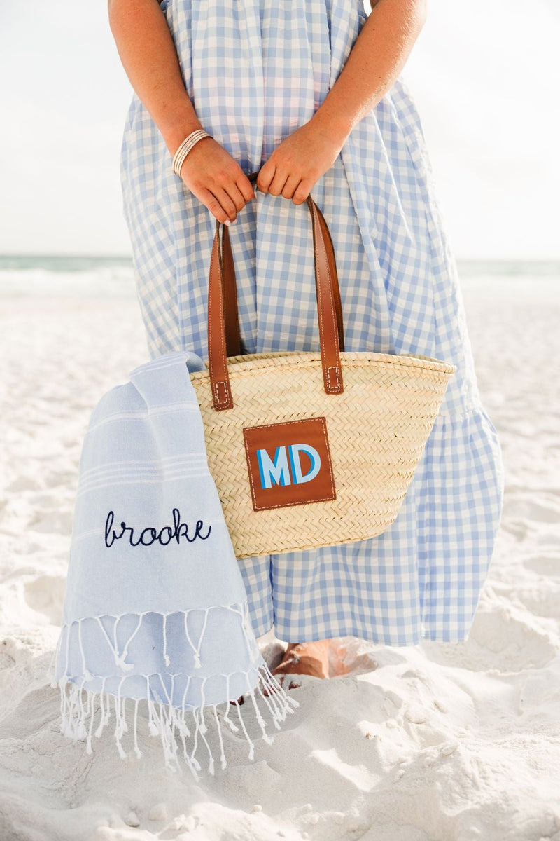 A girl on the beach holds a straw tote with a blue monogram and a towel with an embroidered name in navy thread.