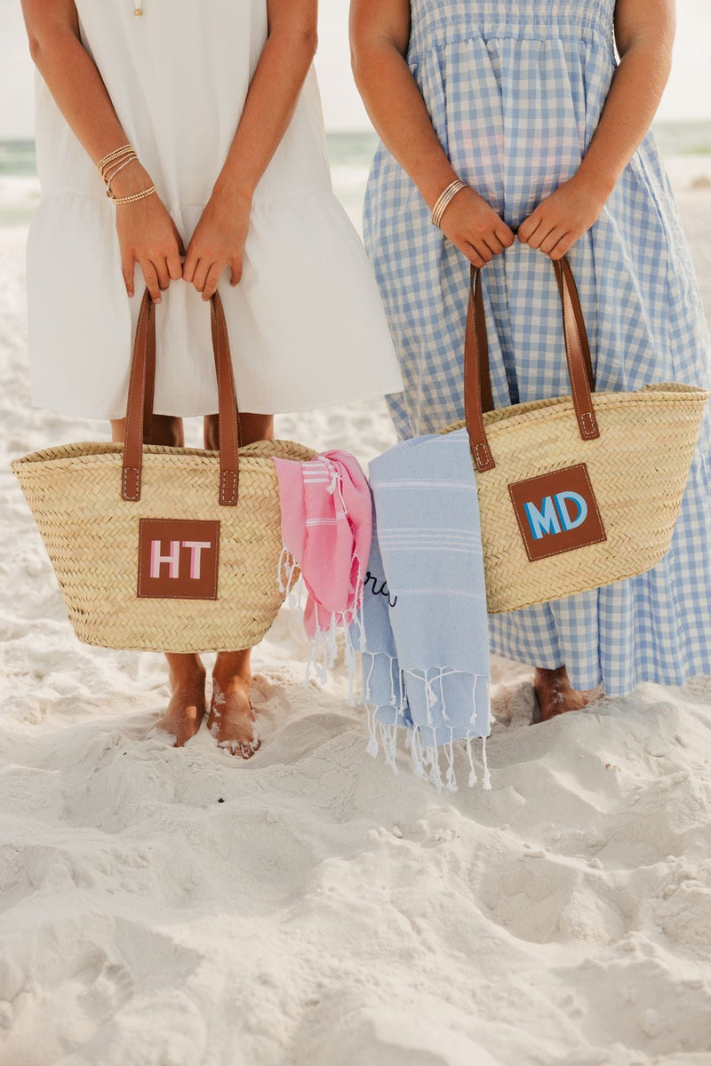 Two girls stand at the beach showing off their straw totes with monograms that match the color of their beach towels