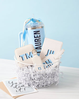 A basket is filled with white leather products and a blue 40 oz tumbler with a blue monogram 