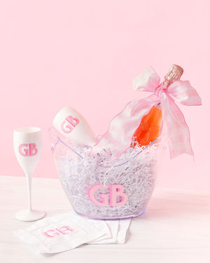 A set of white acrylic champagne flutes, a wide ice bucket and cocktail napkins are customized with a matching pink monogram.